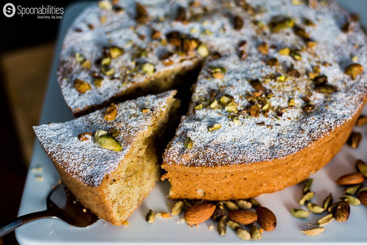 Serving with a cake server a piece of Persian Almond Cardamom Pistachio Cake. The cake is in a white cake stand and garnished with pistachio, almonds & cardamom seeds. Spoonabilities.com