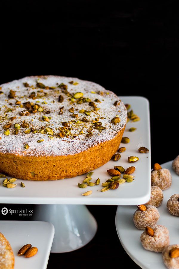Persian Almond Cardamom Pistachio Cake on a white cake stand with powder sugar and garnished with pistachio and cardamom seeds. Next, to another cake stand with Lemony Almond Macaroons. Spoonabilities.com
