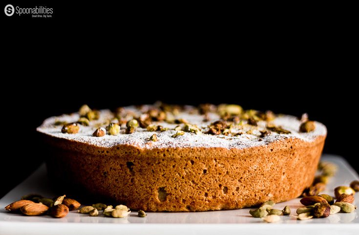 Persian Almond Cardamom Pistachio Cake on a cake platter garnished with pistachios, almonds, cardamom and powder sugar. Spoonabilities.com