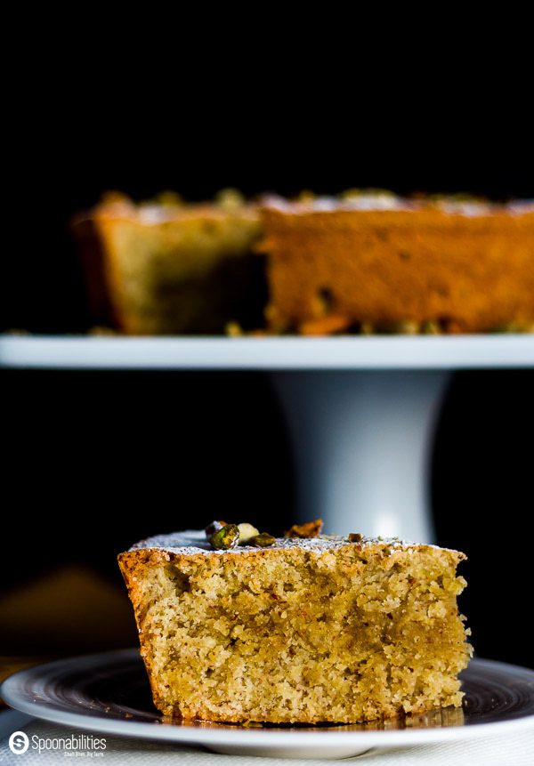 A plated piece of Persian Almond Cardamom Pistachio Cake, with the remaining cake without in the background on a tall white cake stand. Spoonabilities.com