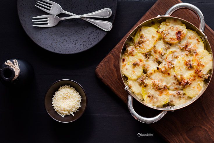 Potato Gratin with leek in the baking pan next to black stone plates and small bowl of grated cheese.
