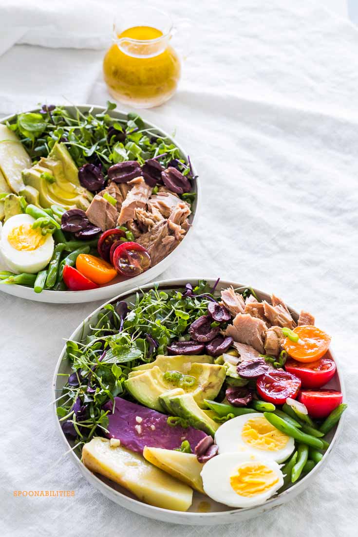 Two bowls of Nicoise Salad on a gray tablecloth with a glass salad dressing pitcher of Dijon mustard vinaigrette