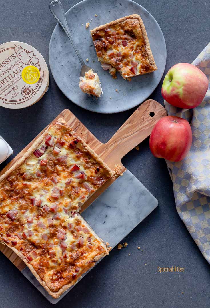 Époisses Ham Apple Tart on a wooden/marble board and a piece of the tart on a round grey plate with a bite on the fork. Two whole pink lady apples, and Époisses cheese in the box. Spoonabilities.com