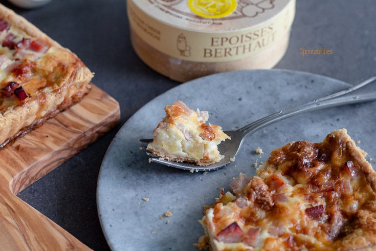Closeup of a piece of Ham Apple Tart on a fork on a round gray plate with the wooden box of the Époisses cheese in the background. Spoonabilities.com