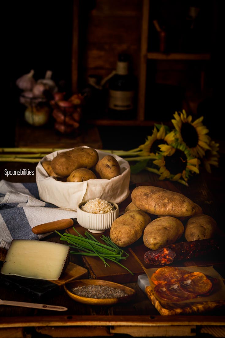 Wooden table filled with Spanish ingredients of chorizo & Lomo Iberico, Manchego cheese & aioli, preparing to make Spanish style baked potatoes. Spoonabilities.com