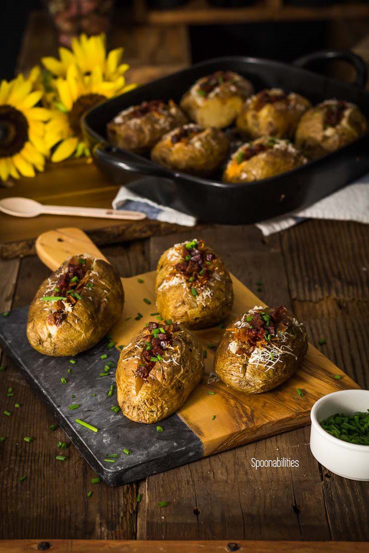 Four Spanish style baked potatoes on a rustic cutting board with six baked potatoes in a pan in the background. Perfect comfort food. Spoonabilities.com