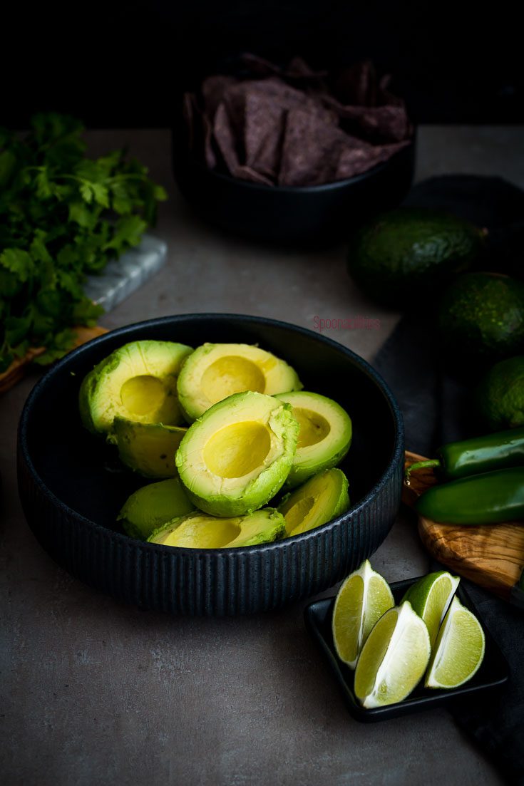 The best guacamole recipe is easy to make. In this photo the avocado meat is in a round black bowl with lime wedges, jalapeño and cilantro. More Mexican recipes and Cinco de Mayo food at Spoonabilities.com