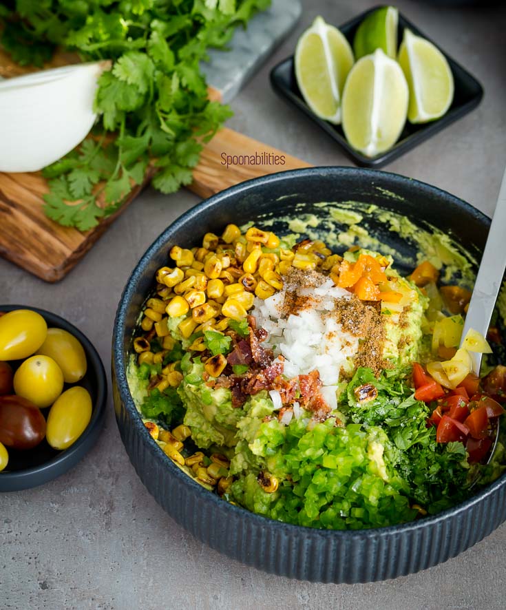 Ingredients for the best guacamole recipe in a black bowl with avocado, lime, cilantro, jalapeño, coriander, cumin, bacon, charred corn. Spoonabilities.com