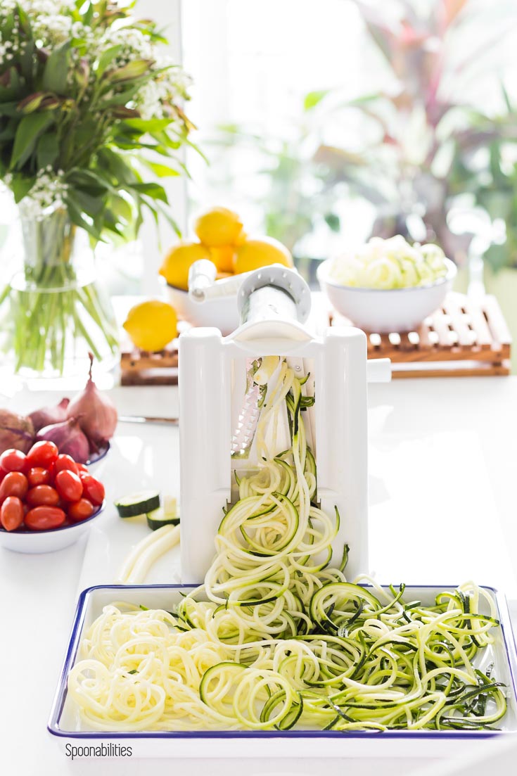 Making Zucchini noodles with the Paderno Spiralizer. Noodles in a white tray with tomatoes, shallots & lemons in the background