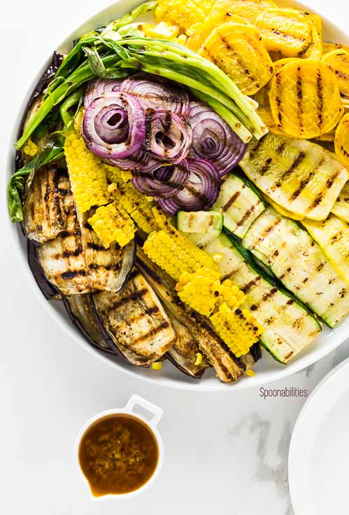 Grilled Summer Vegetables in a round white plate with the Black Olive Vinaigrette in a small white bowl. Spoonabilities.com