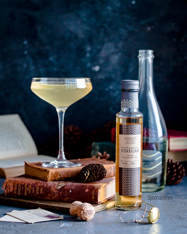 The Gin Prosecco Cocktail is on top of antique books. Next to the cocktail is on of the ingredients, a bottle of white balsamic vinegar. Spoonabilities.com