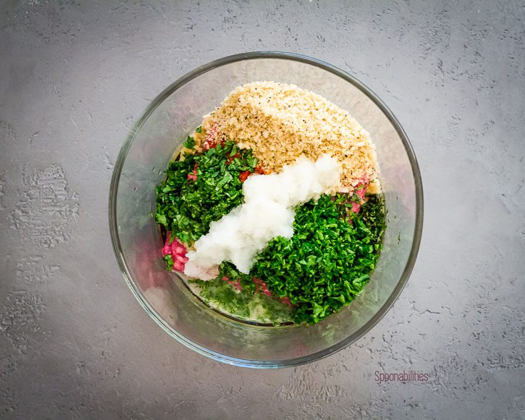 Ingredients in the mixing bowl for the spiced lamb meatballs. In the bowl are parsley, cilantro, mint, breadcrumb, ground lamb, grated onions, egg & spices. Spoonabilities.com