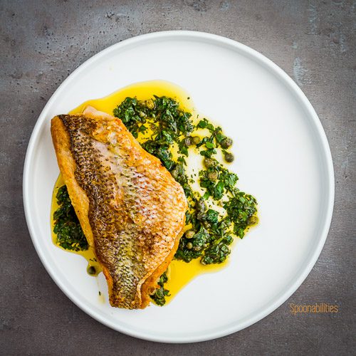 Round white plate with Crispy-Skinned Snapper with Caper Salsa. This salsa has fresh parsley, chili paste, cilantro, olive oil and lemon juice. Spoonabilities.com