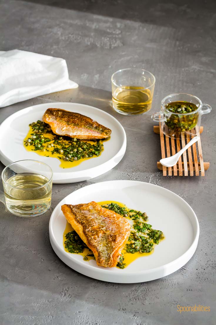 Two round white plates with Crispy Skin Snapper with Caper sauce in a small glass bowl. Two glasses of white wine. Spoonabilities.com