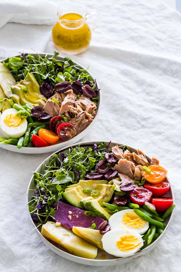Nicoise Salad in two bowls on a gray tablecloth with a glass salad dressing pitcher of Dijon mustard vinaigrette