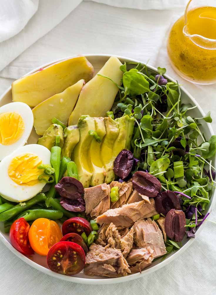 Large bowl of Nicoise Salad with glass pitcher of Dijon mustard vinaigrette, on a gray tablecloth