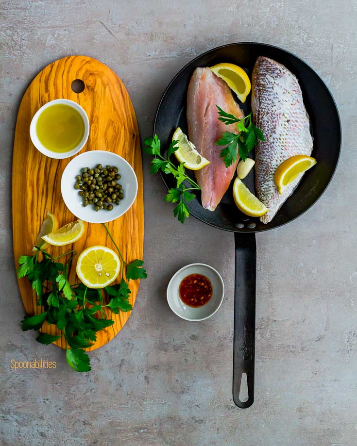 Raw Snapper fish in a fry pan next to a wooden board with capers, lemon, flat parsley, olive oil & Chili paste. Spoonabilities.com