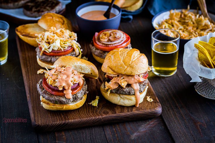 Four burgers presented in a wooden tray on a table with beers, plantain chips, and other food items. Spoonabilities.com