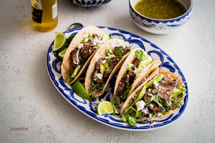 Horizontal photo with four skirt steak tacos in a oval plate and one beer and small bowl in the background. Spoonabilities.com