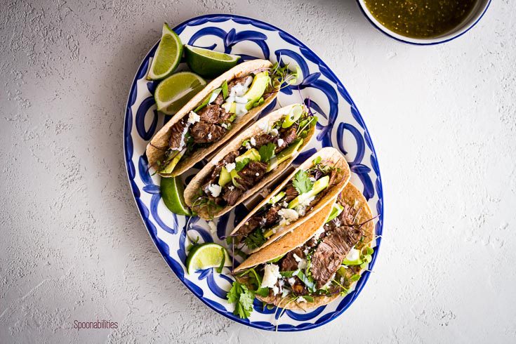 Overhead photo with four Skirt steak tacos in a oval plate and in a small bowl tomatillo salsa. Spoonabilities.com