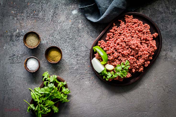 Ground beef in a oval wooden tray in a corner and in the center left three small bowl with seasoning and one small oval wooden tray with cilantro. Spoonabilities.com