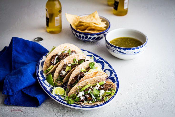 Blue & White oval plate with four Skirt Steak Tacos, one small bowl with tomatillo salsa, another bowl with chips and two beers. Spoonabitlies.com