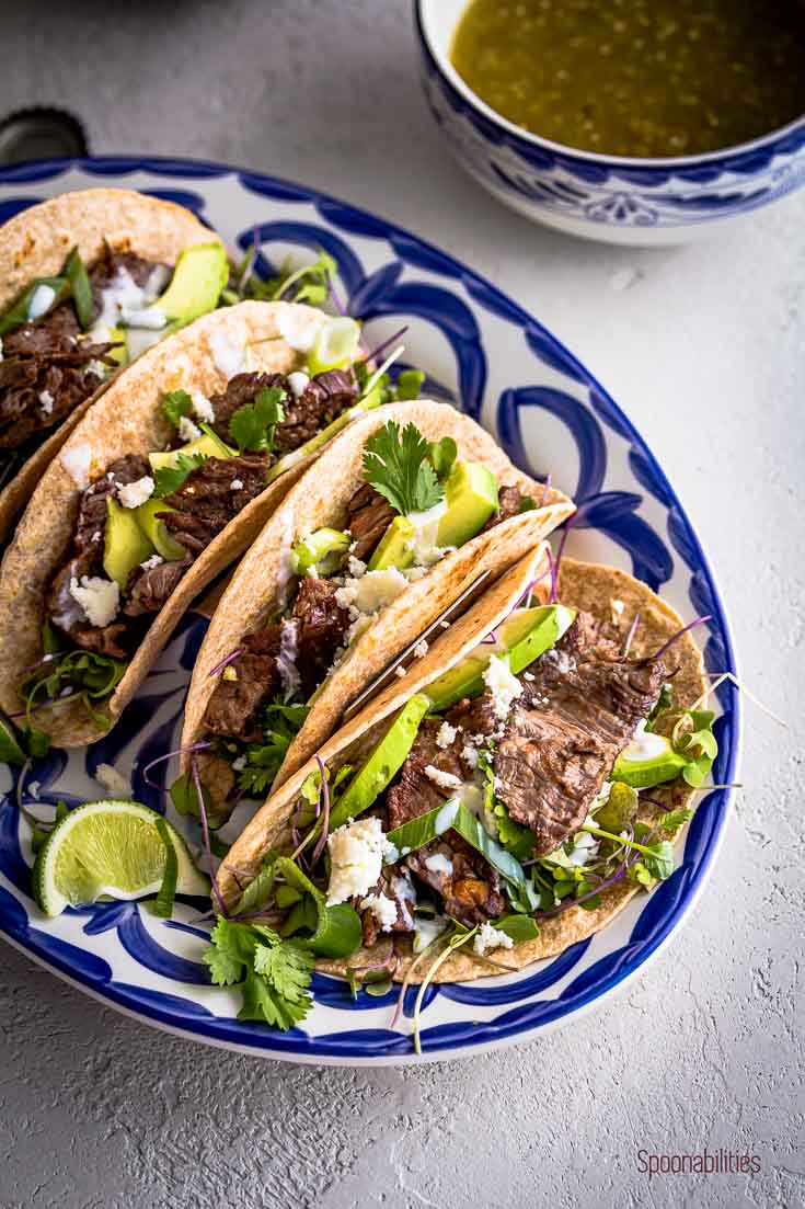 Close up Skirt steak tacos in a white and blue oval plate and in the back a small bowl with tomatillo salsa. Spoonabilities.com