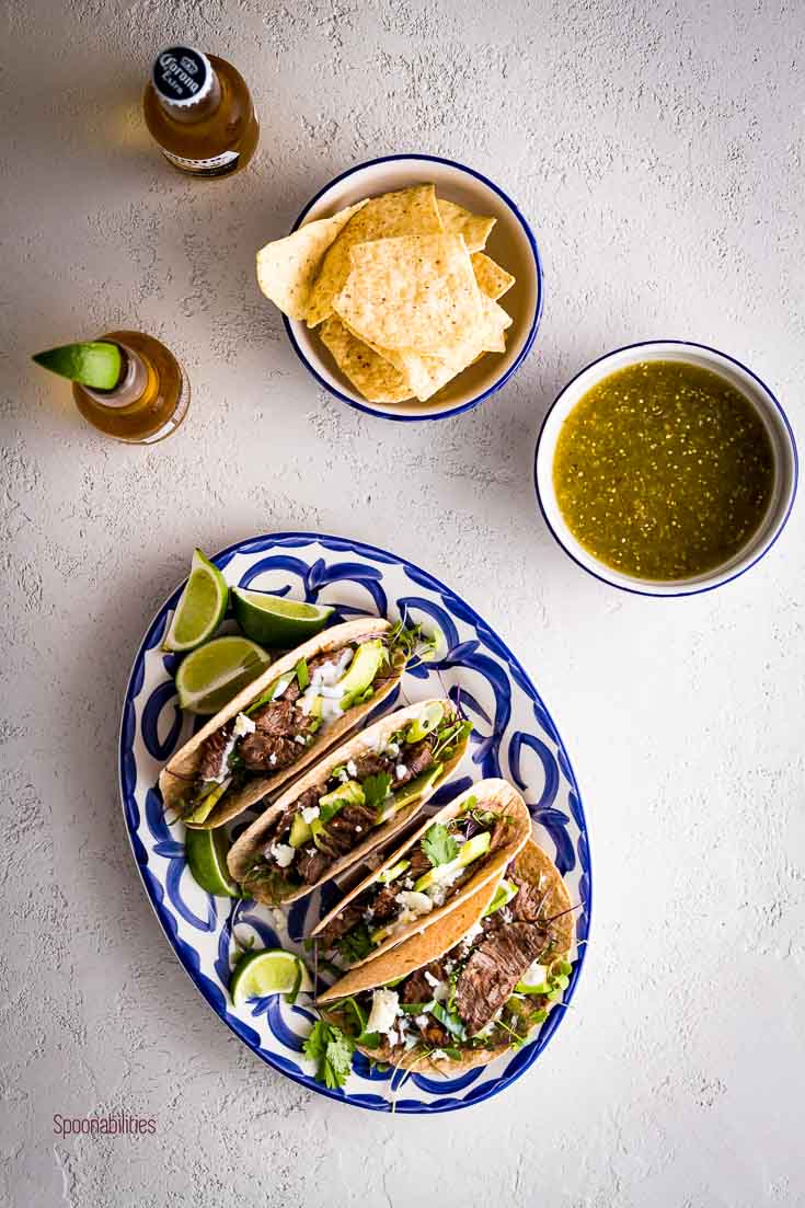 Flat lay photo with an oval serving plate with four skirt steak tacos and two small bowls with chips and tomatillo salsa. Spoonabilities.com