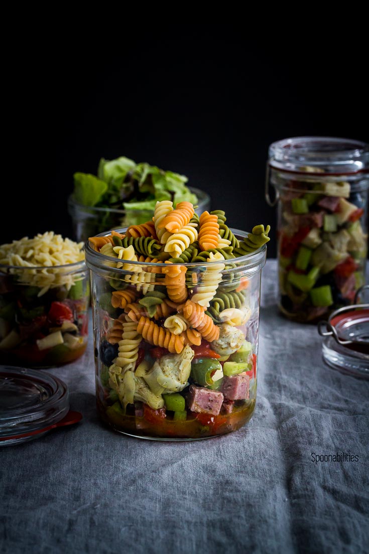 A Jar with Antipasto Pasta Salad and three jars in the background with salad. Spoonabilities.com