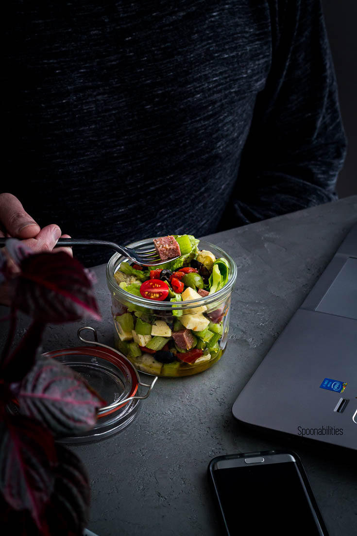 A guy eating salad in a jar with antipasto and lettuce next to his laptop. Spoonabilities.com