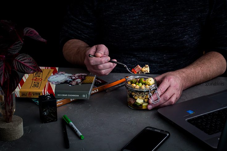 A guy eating salad in a jar on his desk at work with laptop and books around the desk. Spoonabilities.com