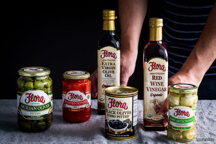 Display of Flora Fine Foods products like black olives, Sicilian olives, roasted red pepper, red wine vinegar, olive oil & baby artichoke. Spoonabilities.com