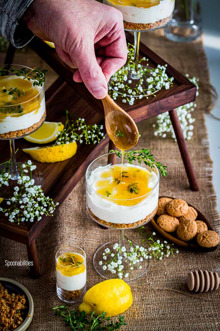 One hand drizzling the Honey Thyme Drizzle over the cheesecake. The cheesecake is in a cocktail glass. Spoonabilities.com