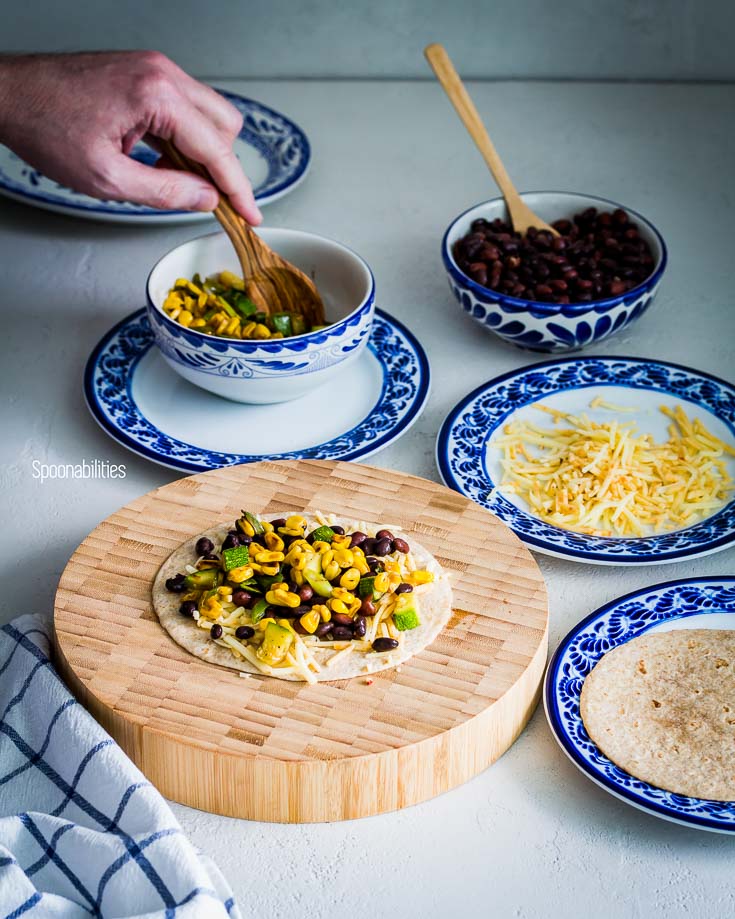 Prepping the vegetarian quesadillas. Open face tortilla on a wooden round board with cheese, black beans, charred corn & zucchini and four plates around with the different ingredients. Spoonabilities.com