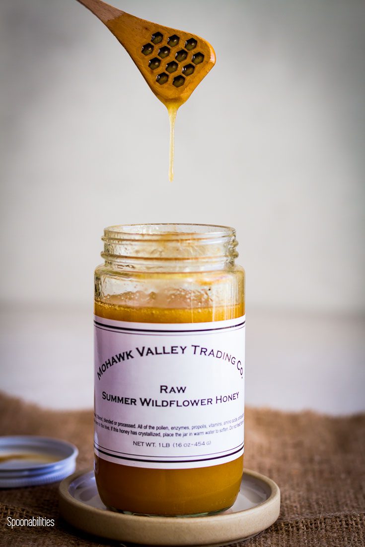 Open jar of Raw Summer Wildflower Honey and one hand with a honey dipper in the air dripping honey into the jar. Spoonabilities.com