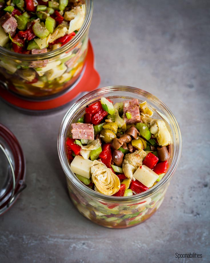 Close up of a salad in a jar with baby artichokes, black olives, green olives, provolone, hard salami, celery with Italian dressing. Spoonabilities.com