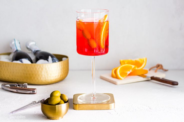 Tall cocktail glass in the center with the Italian drink Aperol Spritz with green olives. In front of the glass a small golden bowl with Greek olives and in the background two bottles of prosecco and orange slices. Spoonabilities.com