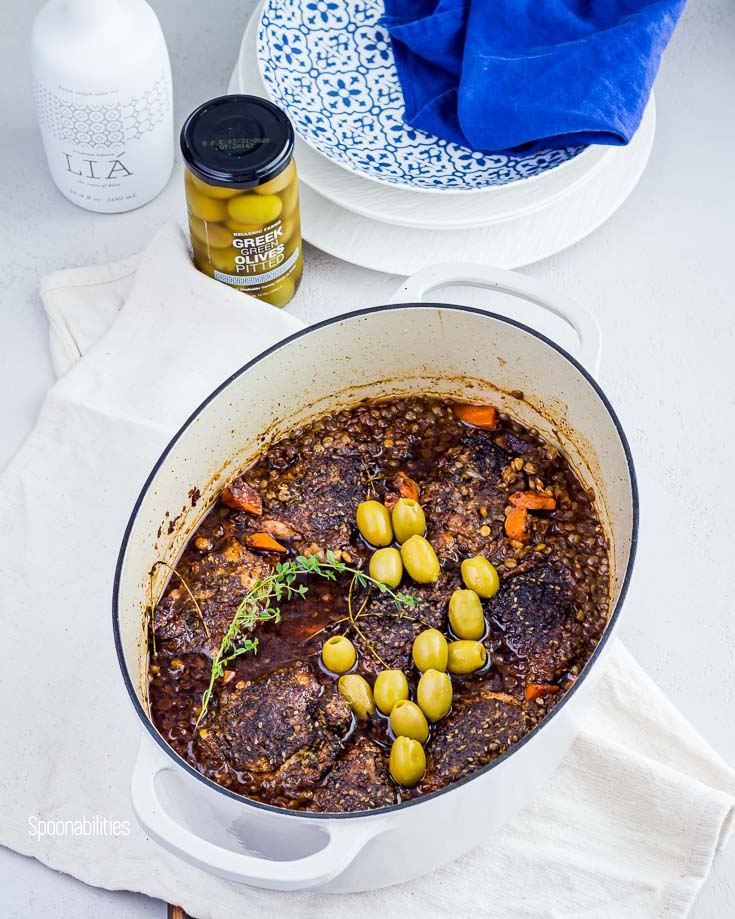 Lodge white oval Dutch oven pot with chicken thighs, lentils, carrots with Greek olives. In the background a jar of Pitted green olives and Greek premium Lia extra virgin olive oil. Spoonabilities.com