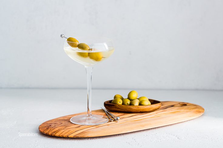 A Cocktail glass on top of a wooden board with the drink dirty martini and garnished with three olives in a metal pick. In the background pitted green olives in a small oval wooden bowl. Spoonabilities.com