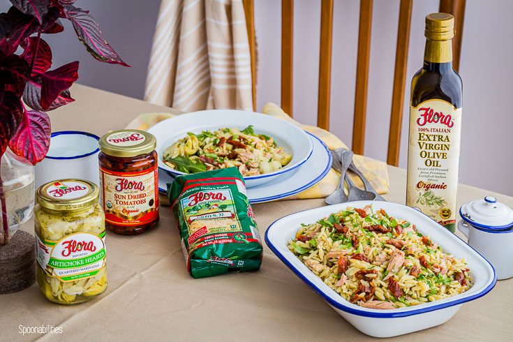 Dining table with a white serving dish with the orzo tuna salad and a round white plate in the back with salad. And, around the table the Flora Fine Food products like artichoke hearts, sun-dried tomatoes, orzo pasta and olive oil. Spoonabilities.com