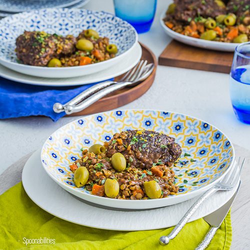 Braised Mediterranean Chicken Thighs in a round pasta plate with olives and lentils and in the background another plate and a serving platter. Spoonabilities.com