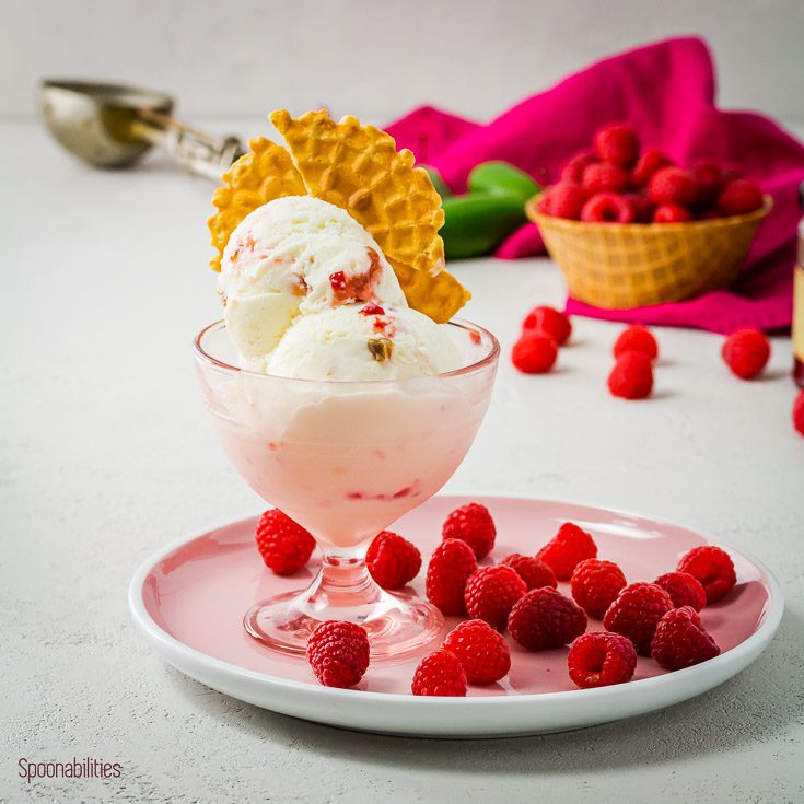 Light pink ice cream bowl on top of a pink round plate with Raspberry around the bowl. In the ice cream bowl you have two big Raspberry Jalapeño Ice Cream with a Pizzele as a garnish. Spoonabilities.com