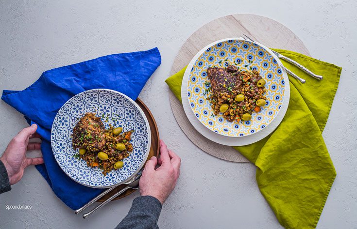 overhead photo with Two round bowl plates with chicken, lentils & olives with two hands holding the bowl in the left side. Spoonabilities.com