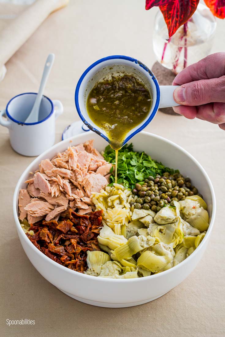 White mixing bowl with orzo, artichoke, sun-dried tomatoes, tuna, chopped parsley, capers. Pouring over the salad the Lemony Dijon Mustard Vinaigrette. Spoonabilities.com