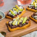 Four Avocado & Black Bean Crostini recipe on a wooden board. Spoonabilities.com