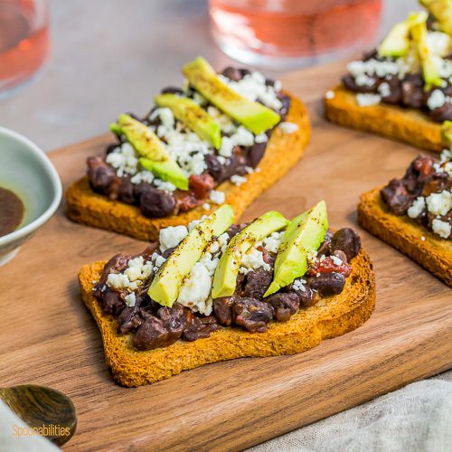 Four Avocado & Black Bean Crostini recipe on a wooden board. Spoonabilities.com