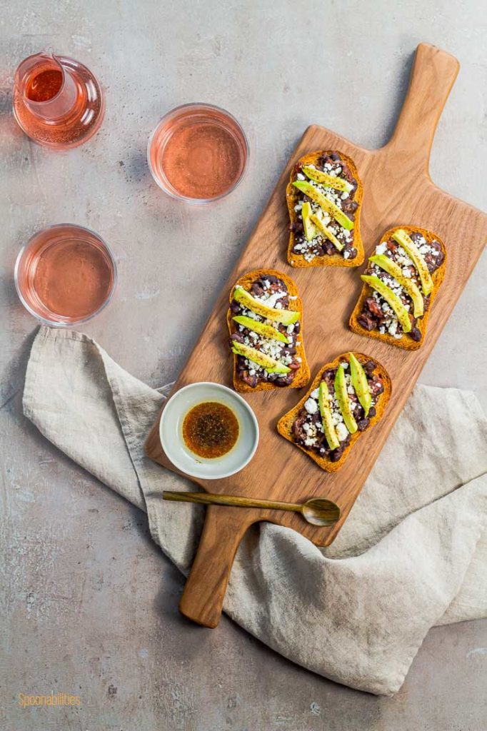 Flat lay photo of four crostini in a wooden board with a small sauce bowl in the bottom left side and a glass of rose wine on the left side. Spoonabilities.com