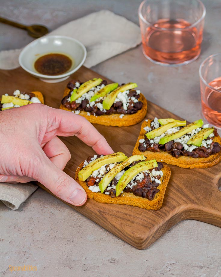 Close up with one hand reaching the black bean crostini from the wooden board. Spoonabilities.com
