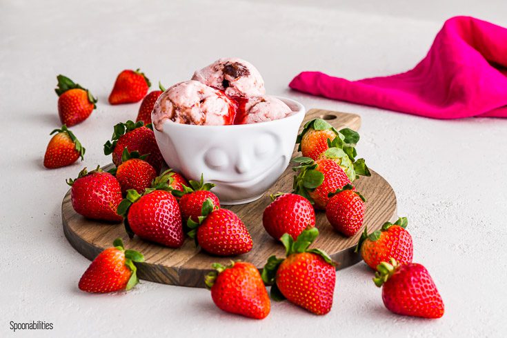 Strawberry ice cream in a white bowl with a smiling face on top of a wooden serving board with fresh strawberry. Spoonabilities.com