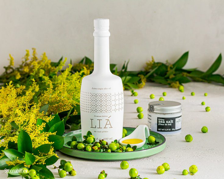 LIÁ premium extra virgin olive oil on top of a green round plate with flowers in the left side and a jar of sea salt fleur de sel by Sal odyssey. Available at Spoonabilities.com